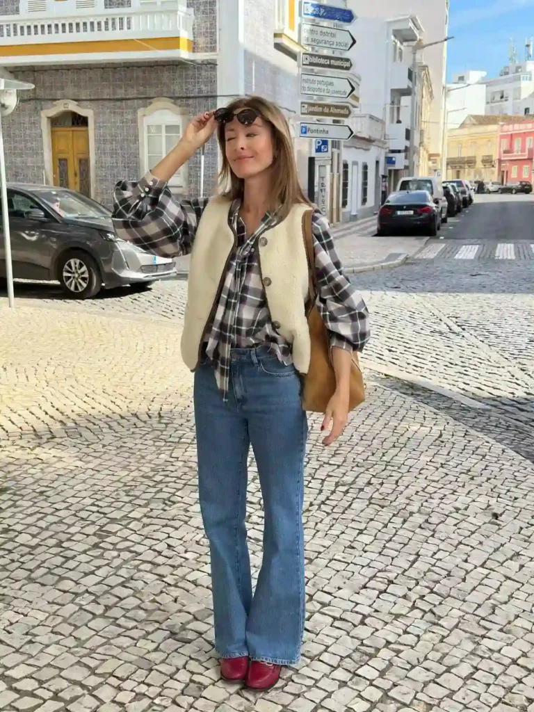 Fashionable woman in plaid shirt and flared jeans enjoying a sunny day on a cobblestone street.