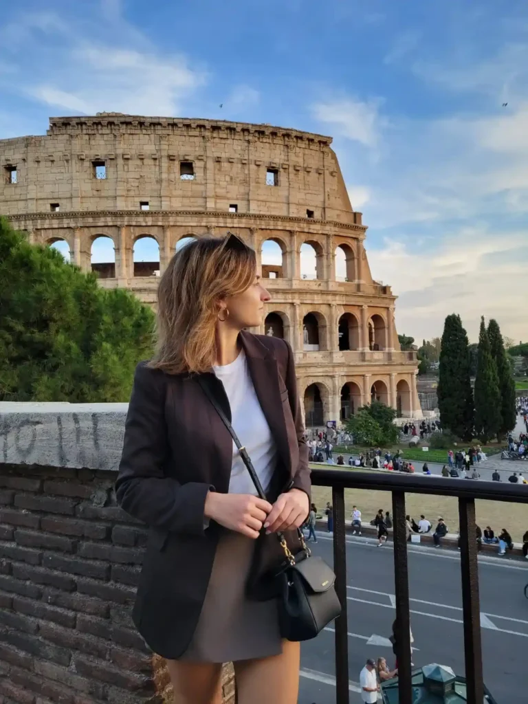 Young woman in a blazer poses near the Colosseum in Rome, surrounded by greenery and a bustling crowd.