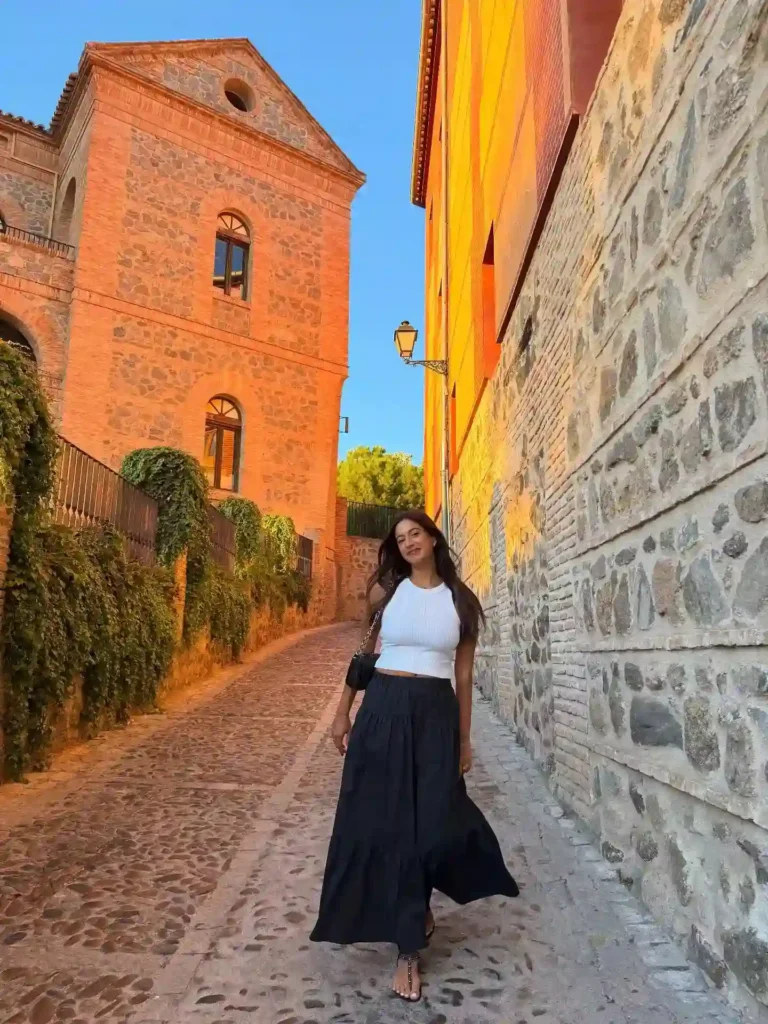 Woman in a stylish outfit walking along a picturesque cobblestone street lined with historic buildings and greenery.
