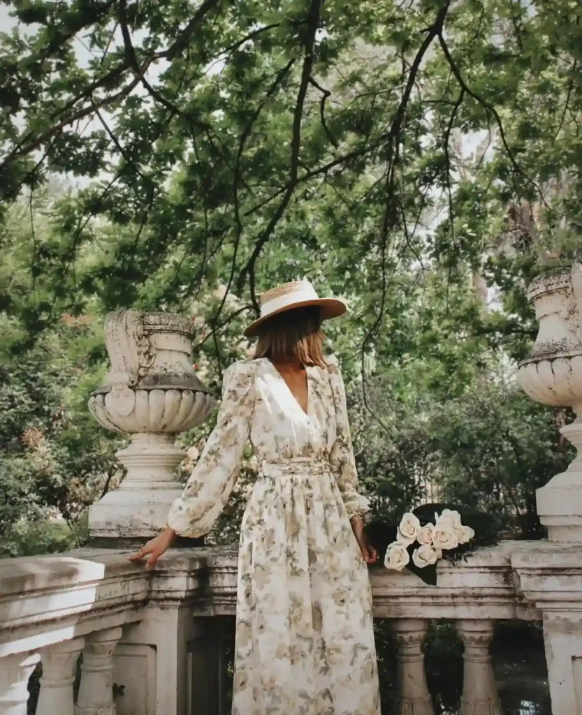 Woman in a floral dress and hat stands by a garden balcony, surrounded by lush greenery and roses. Elegant outdoor scene.