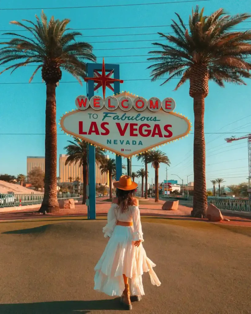 Woman in a flowing white dress and hat walking towards the iconic Welcome to Las Vegas sign, surrounded by palm trees.