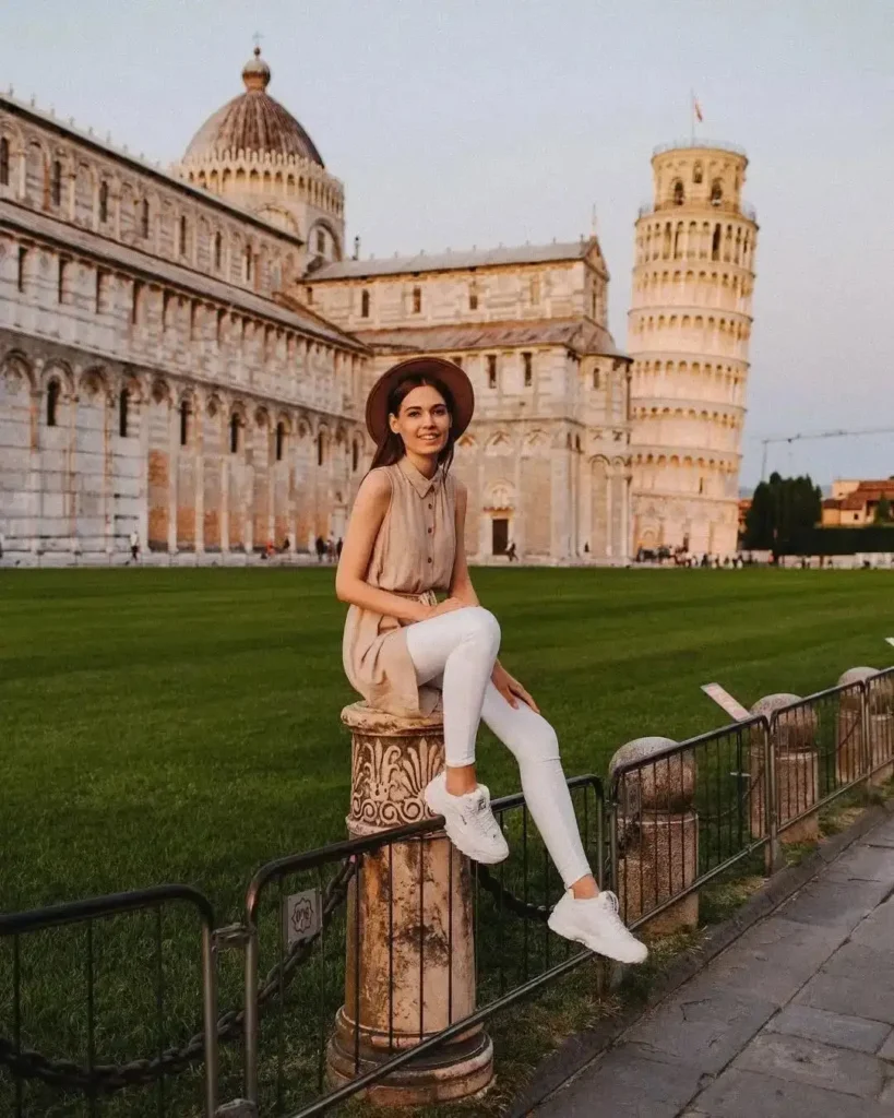Young woman poses on a stone pillar in front of the Leaning Tower of Pisa and historic buildings during sunset.