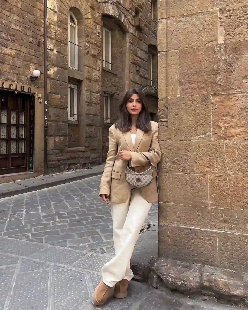 A woman in a stylish beige blazer and cream pants stands against a stone wall in a quaint city street.