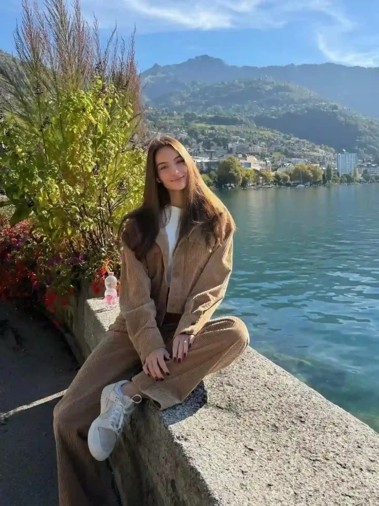 Young woman in casual outfit sitting by a lake, surrounded by greenery and mountains on a sunny day.