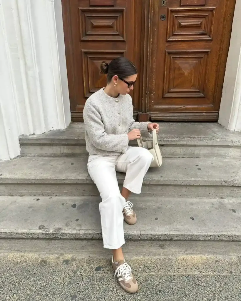 Woman in stylish outfit sitting on steps near a wooden door, showcasing casual chic fashion with sneakers and a handbag.