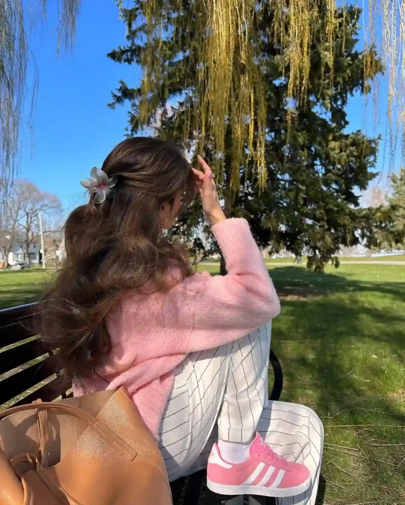 Young woman in a pink sweater sitting on a bench, enjoying a sunny day under trees in a park setting.