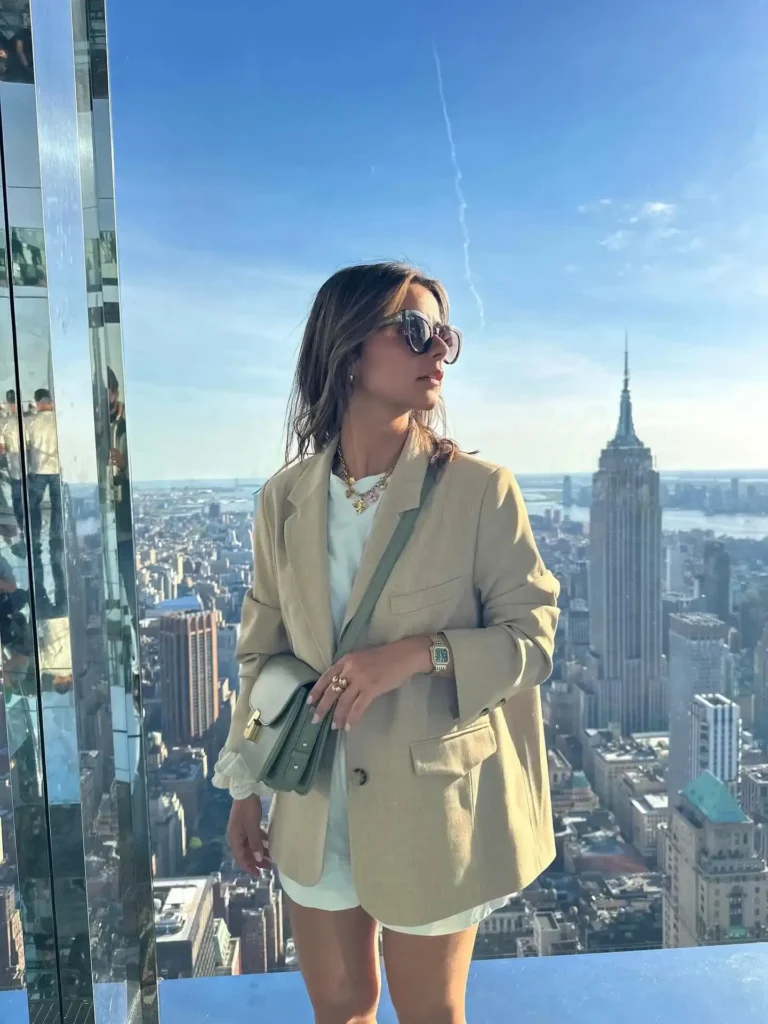 Woman in a stylish blazer and sunglasses poses against a stunning New York City skyline, featuring the Empire State Building.
