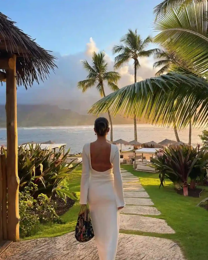 Woman in a white dress strolling along a tropical path, with palm trees and ocean in the background at sunset.