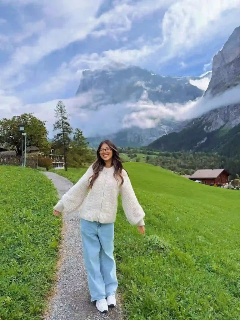 Young woman in cozy sweater smiling on a scenic mountain path, surrounded by lush greenery and dramatic clouds.