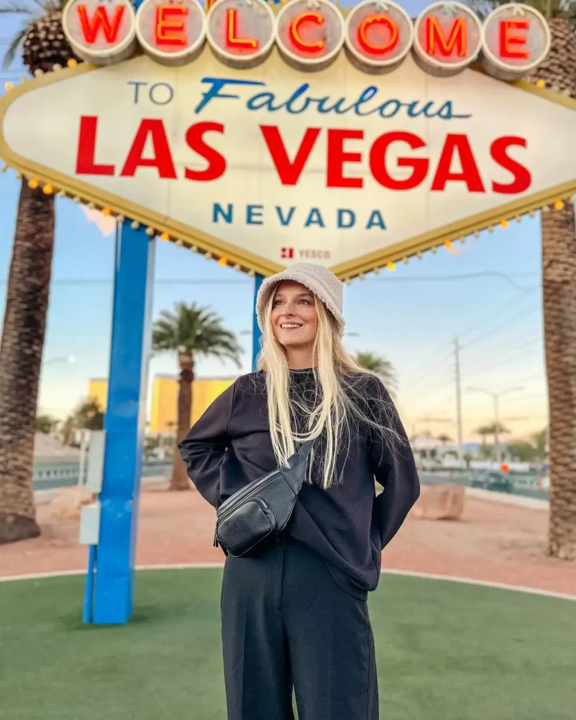 Young woman in casual attire stands in front of the iconic Welcome to Fabulous Las Vegas sign at sunset.