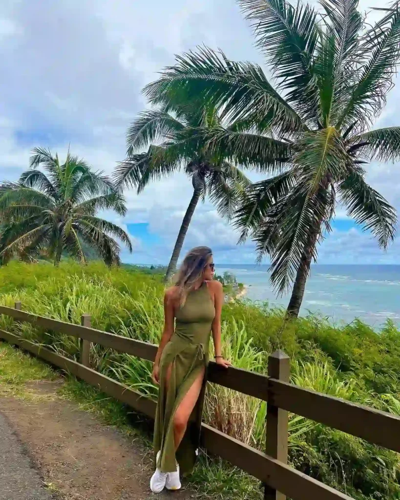 Woman in a green dress poses by a wooden fence with palm trees and ocean views in a tropical landscape.