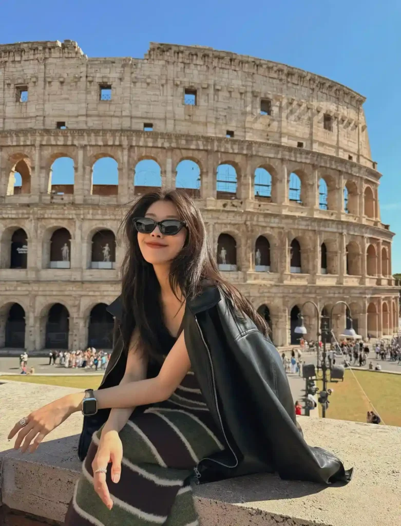 Stylish woman in sunglasses poses near the Colosseum in Rome, showcasing iconic architecture against a clear blue sky.