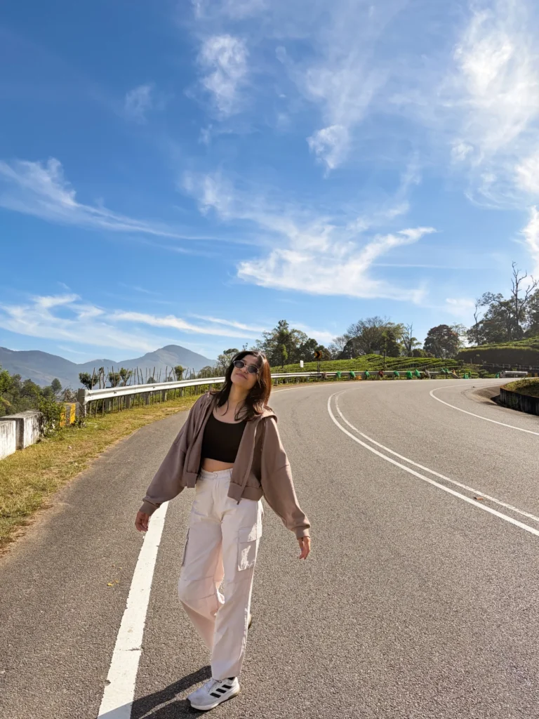 A woman casually walks along a scenic road under a bright blue sky, enjoying a sunny day in nature.