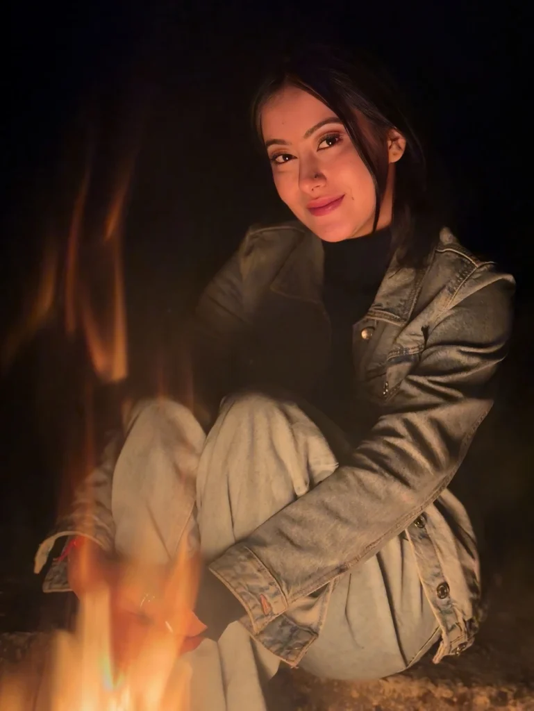 Young woman sitting by a fire, smiling warmly, wearing a denim jacket and cozy jeans, illuminated in the dark.