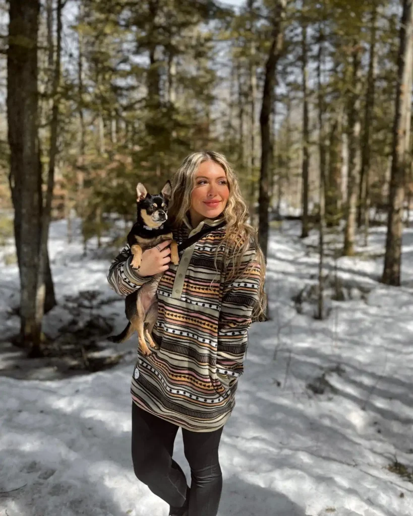 Woman in a striped sweater holds a small dog in a snowy forest, surrounded by tall trees and winter scenery.