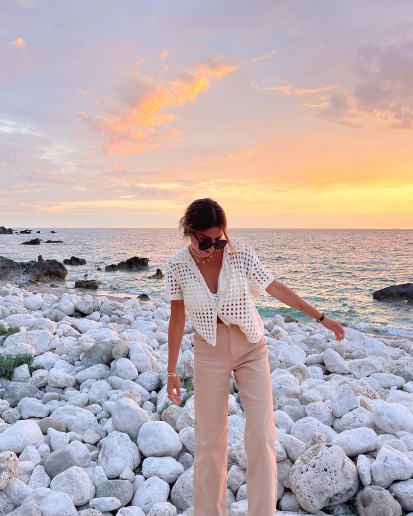 Woman in a white crochet top and beige pants standing on rocky beach at sunset, enjoying the serene ocean view.
