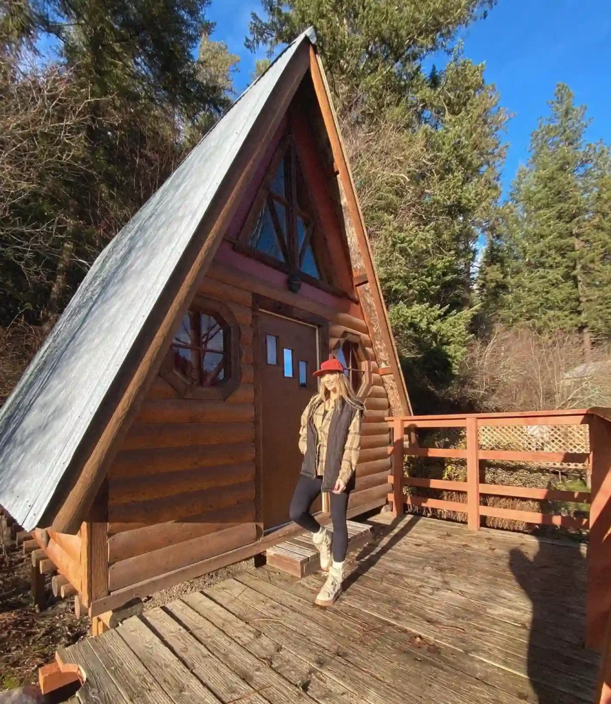 Charming A-frame cabin in a wooded area, featuring a wooden deck and a woman in stylish outdoor attire.