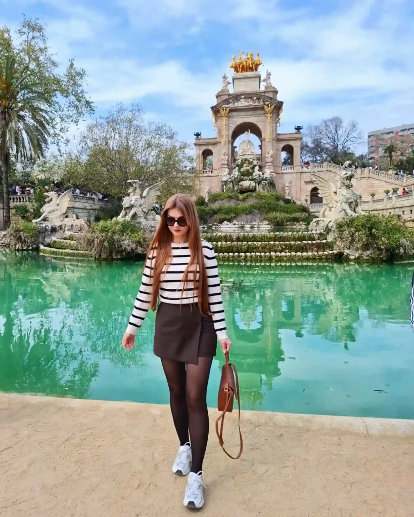 Young woman in sunglasses walking by a green lake, with a grand golden architectural structure in the background.