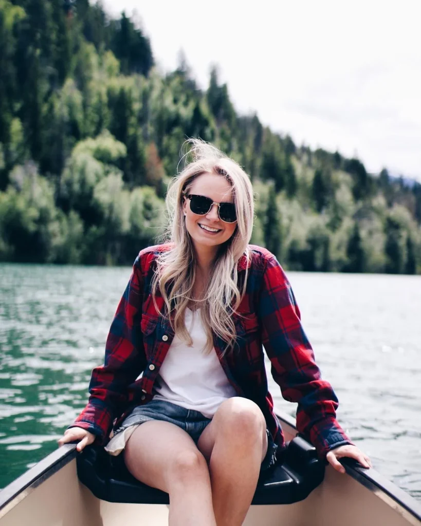 Smiling woman in sunglasses and plaid shirt sits in a canoe on a serene lake surrounded by lush greenery.