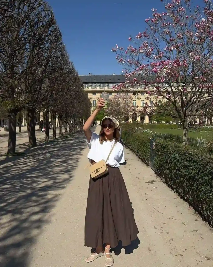 A woman in a stylish outfit enjoys a sunny day in a park lined with trees and blooming flowers.