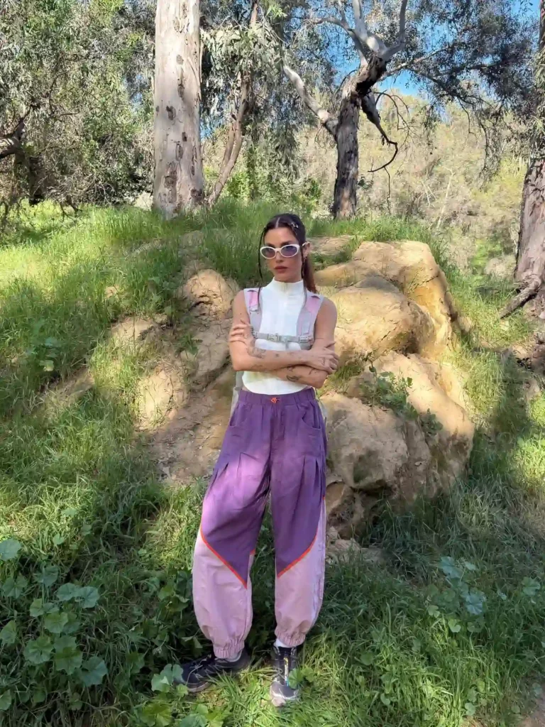 A person in a trendy outfit poses confidently outdoors among trees and rocks in a lush green setting.