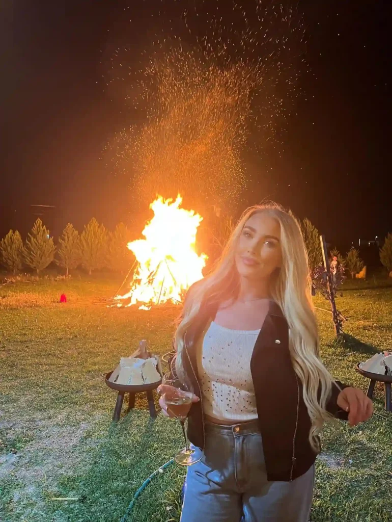 A woman with long hair poses with a drink beside a large bonfire in a nighttime outdoor setting.