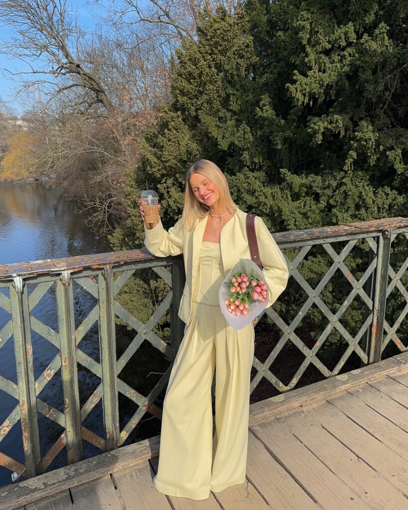 Cheerful woman in pastel yellow outfit holding coffee and flowers on a bridge by a river, enjoying a sunny day.