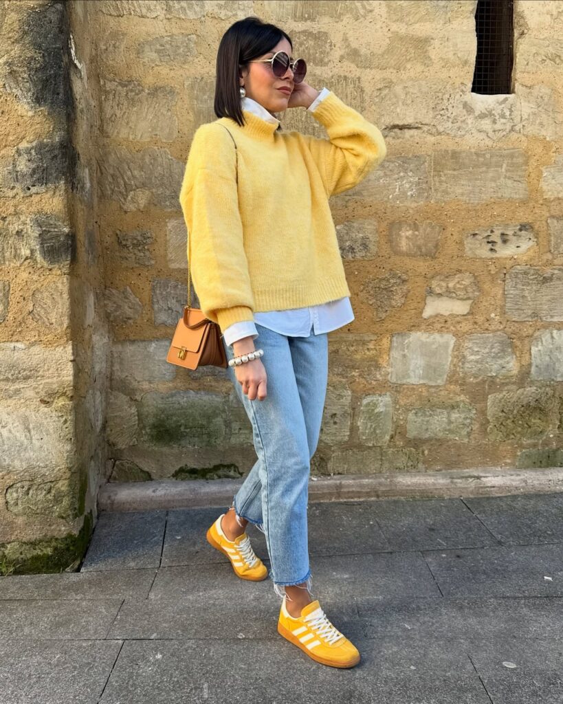 Woman in a yellow sweater and jeans, styled with sunglasses and yellow sneakers, posing against a stone wall.