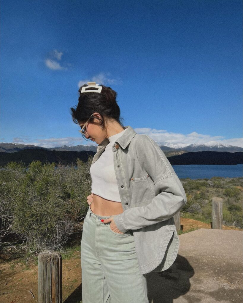 Young woman in a gray shirt and pants stands by a lake, with mountains and blue sky in the background.
