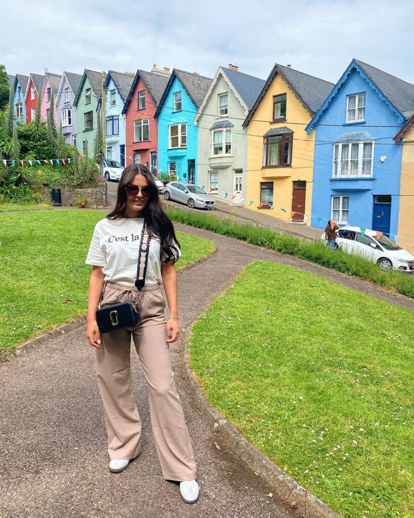 Young woman in stylish outfit standing in front of colorful houses on a sunny day, surrounded by green grass.