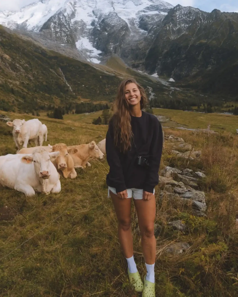 Smiling woman standing in a scenic mountain pasture with cows, showcasing nature's beauty and tranquility.