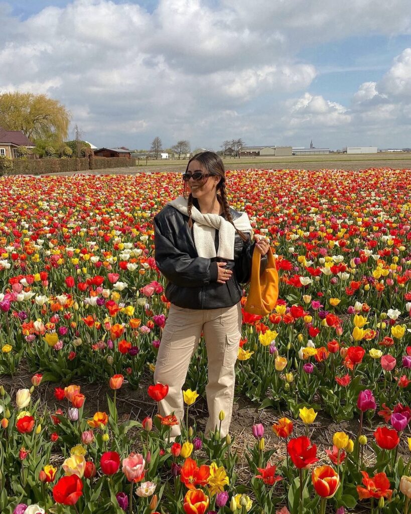 Woman in sunglasses poses with an orange bag in a vibrant tulip field under a blue sky with fluffy clouds.