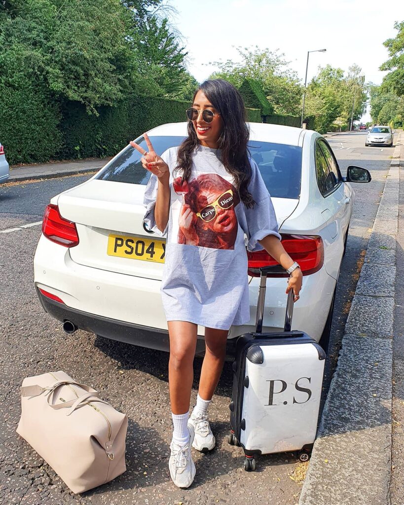 Young woman in stylish outfit posing with peace sign beside car and luggage, ready for a fun day out or travel adventure.