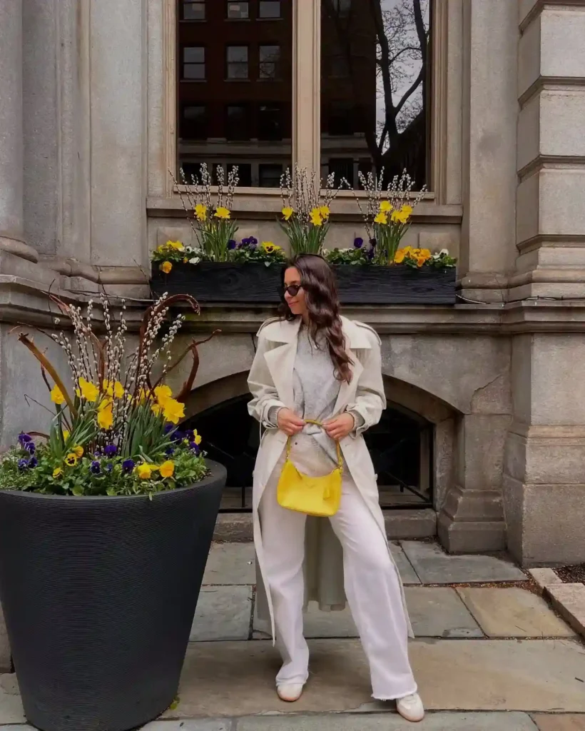 A stylish woman in a cream coat and yellow handbag poses beside vibrant spring flowers by a historic building.