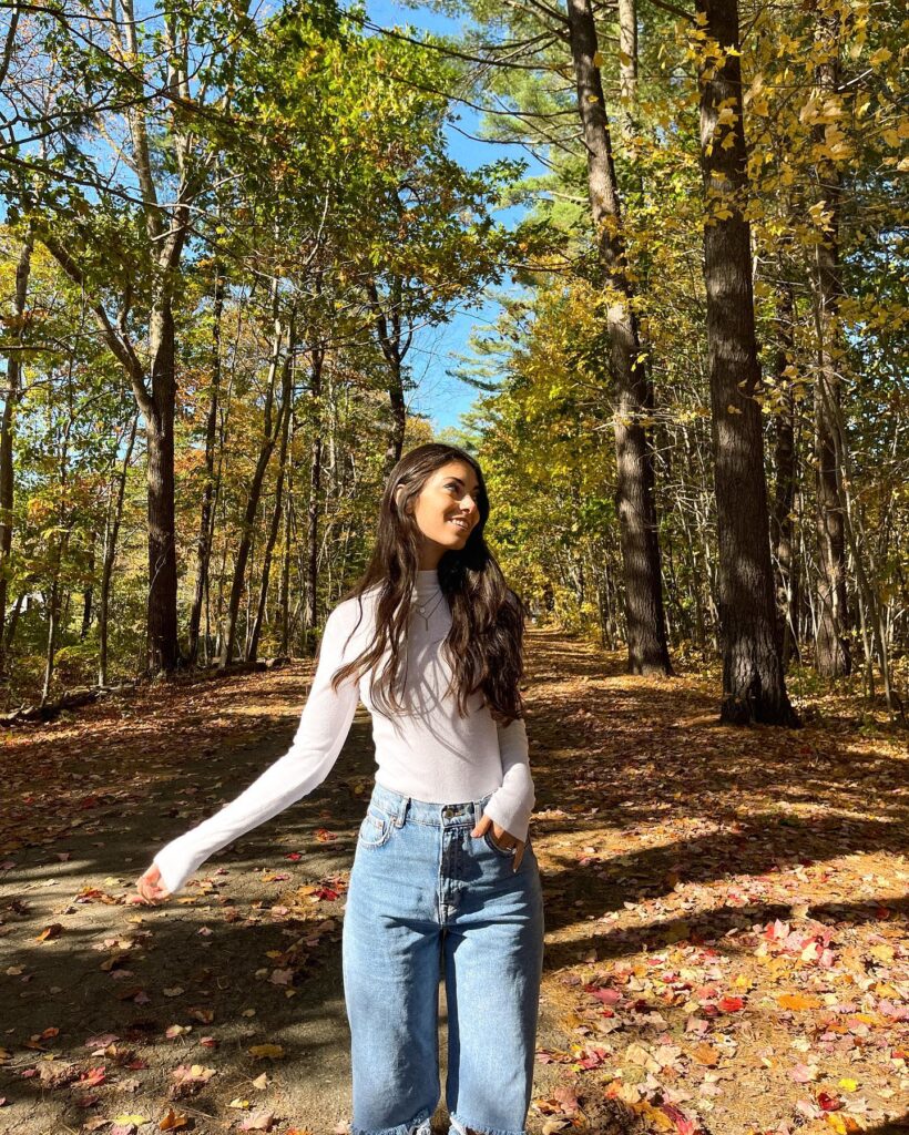 Young woman in a white long-sleeve shirt and denim jeans walking on a forest path covered in autumn leaves.