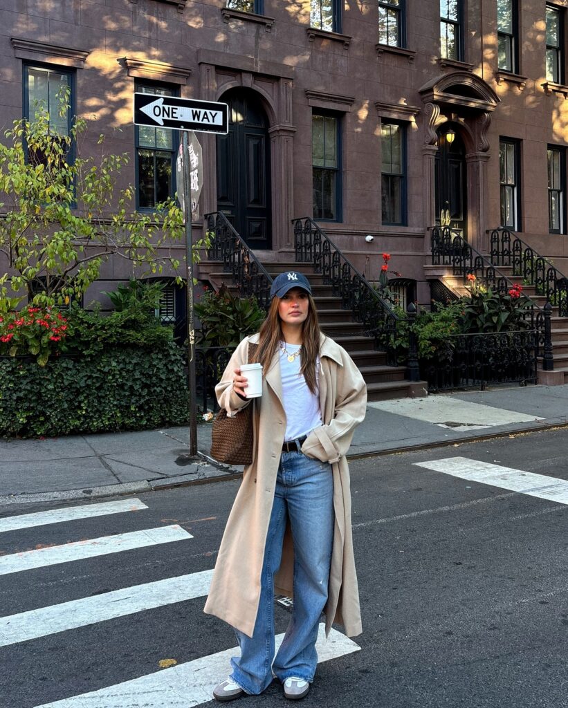A woman in a beige trench coat and baseball cap holds a coffee cup, standing on a city street with brownstone buildings.