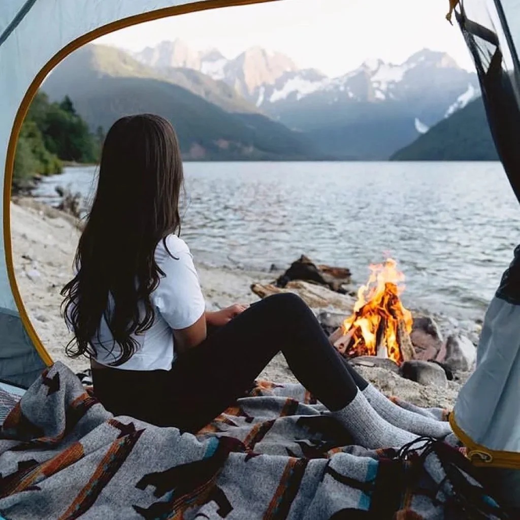 A serene lakeside camping scene with a person relaxing by a campfire and mountains in the background.