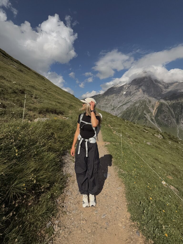 Person hiking on a rugged trail surrounded by mountains and greenery under a partly cloudy sky.
