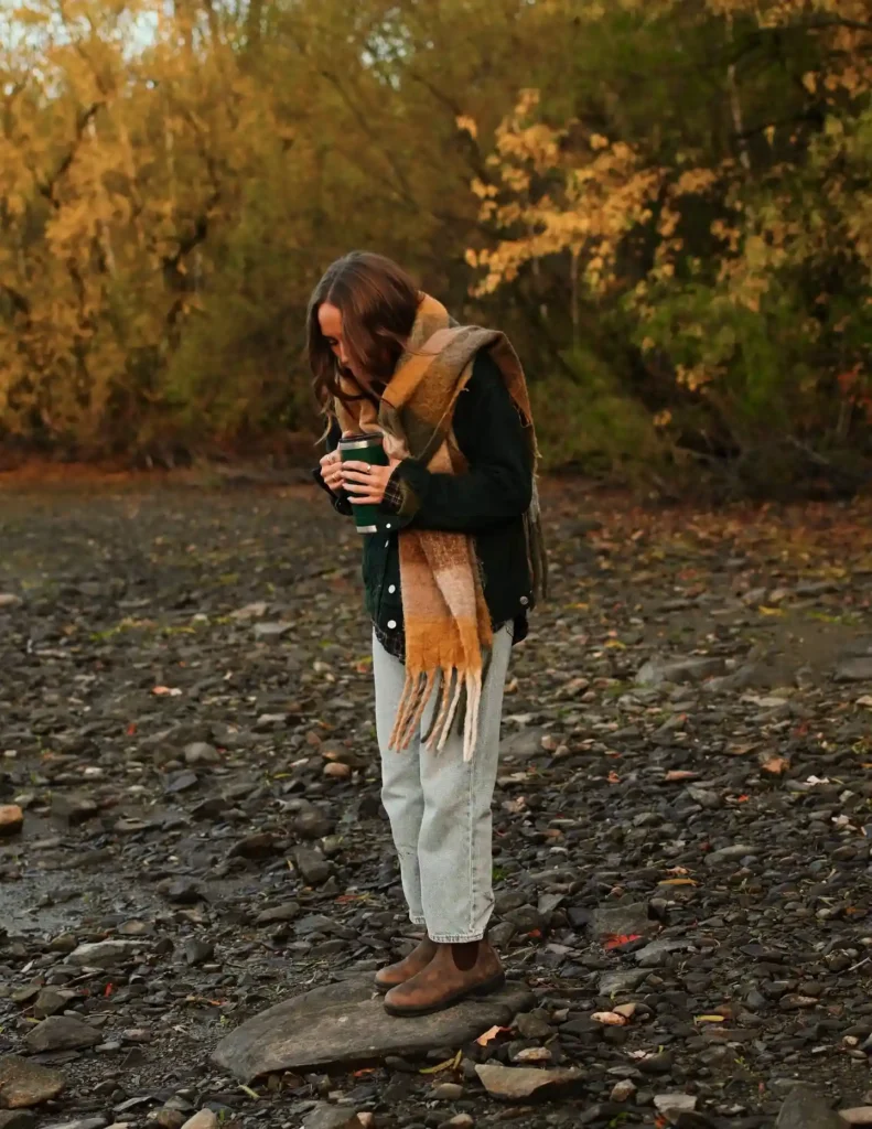Woman in a cozy scarf stands on a rocky shore, enjoying a warm drink amidst autumn foliage. Perfect for fall inspiration.
