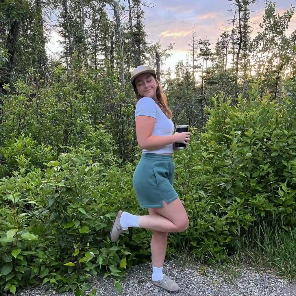 Young woman in a blue outfit poses playfully outdoors, holding a cup amidst lush greenery and a sunset backdrop.