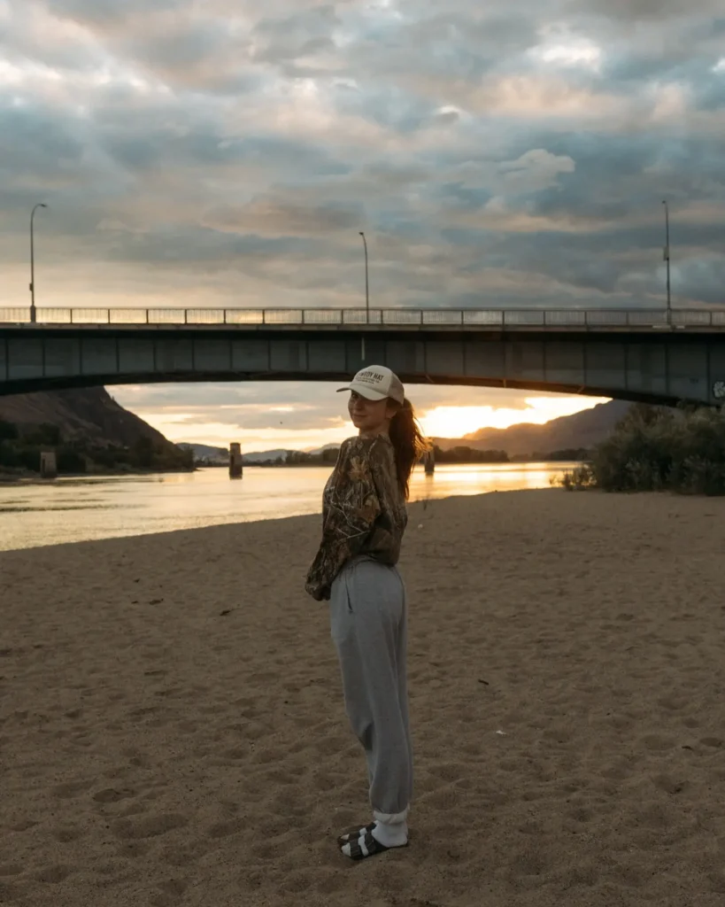 Young woman in camo shirt and sweatpants standing on sandy beach at sunset under a bridge, enjoying nature's beauty.