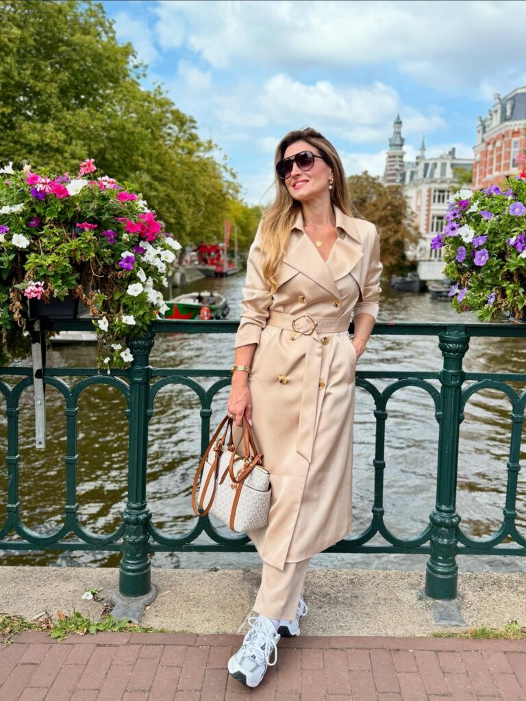 Woman in a stylish beige trench coat poses by a canal, surrounded by flowers and historical buildings on a sunny day.