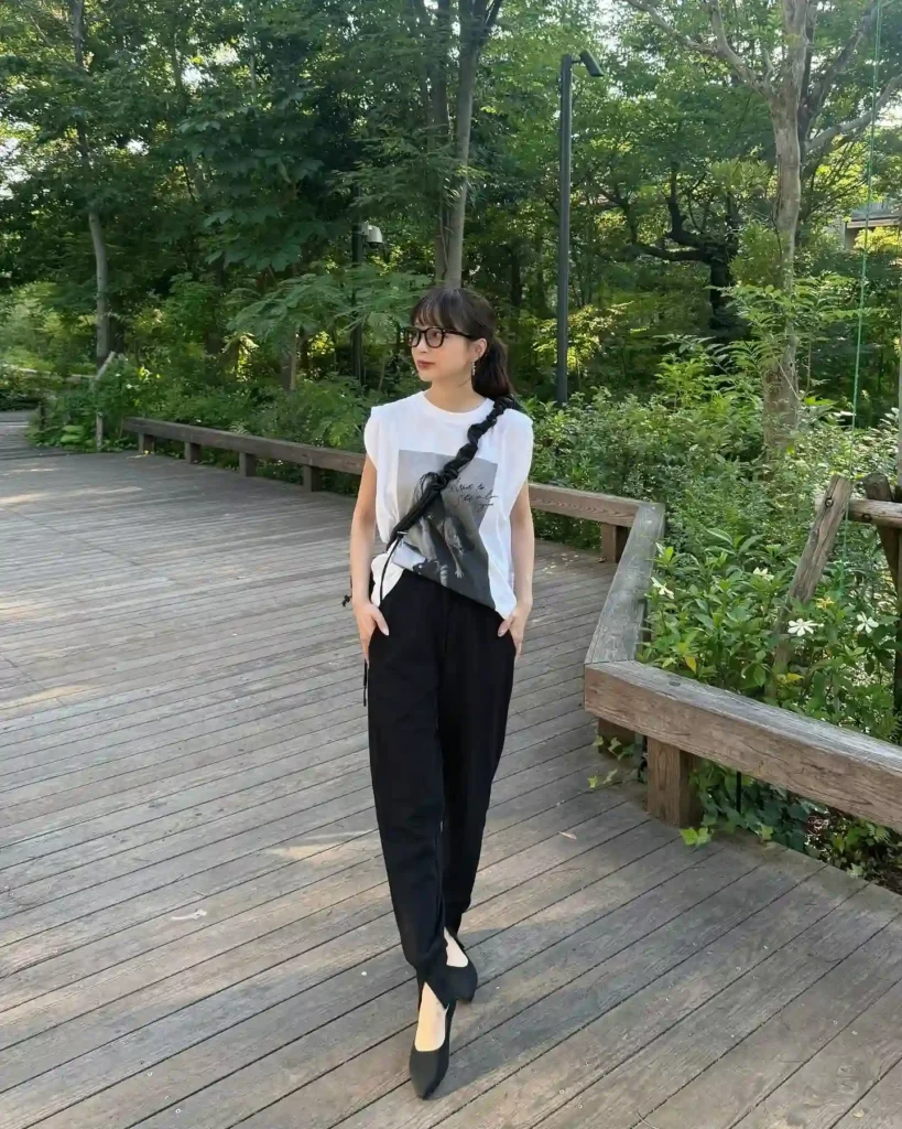 Young woman in stylish black pants and graphic t-shirt strolling along a wooden path in a lush green park.