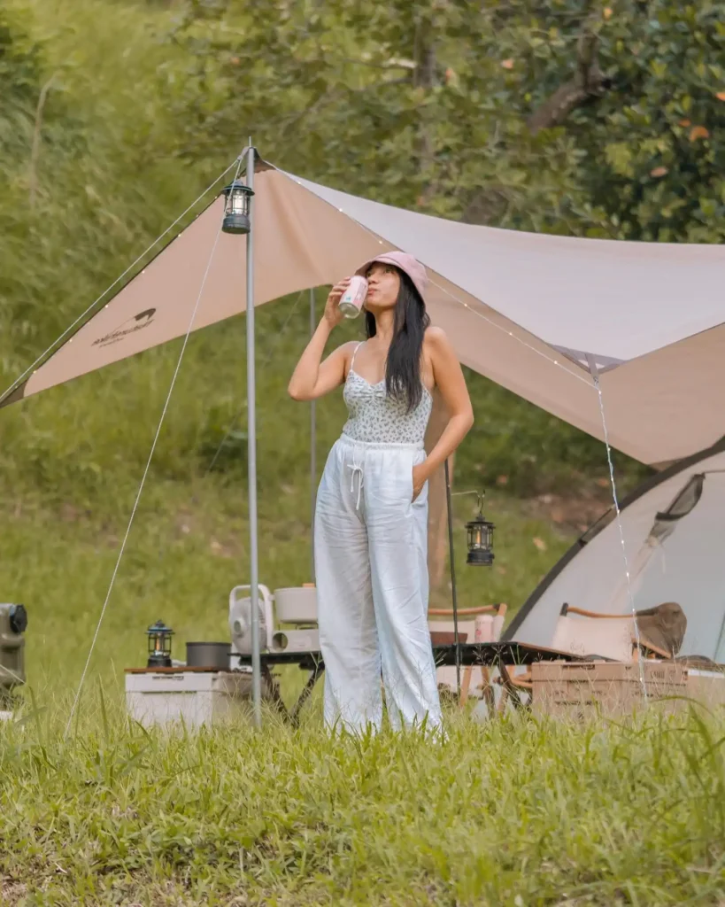 Woman enjoying a drink while camping outdoors under a canopy, with a tent and camping gear in a lush green setting.