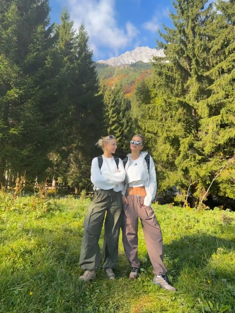 Two young women in stylish outdoor outfits pose in a lush green forest with mountains in the background.