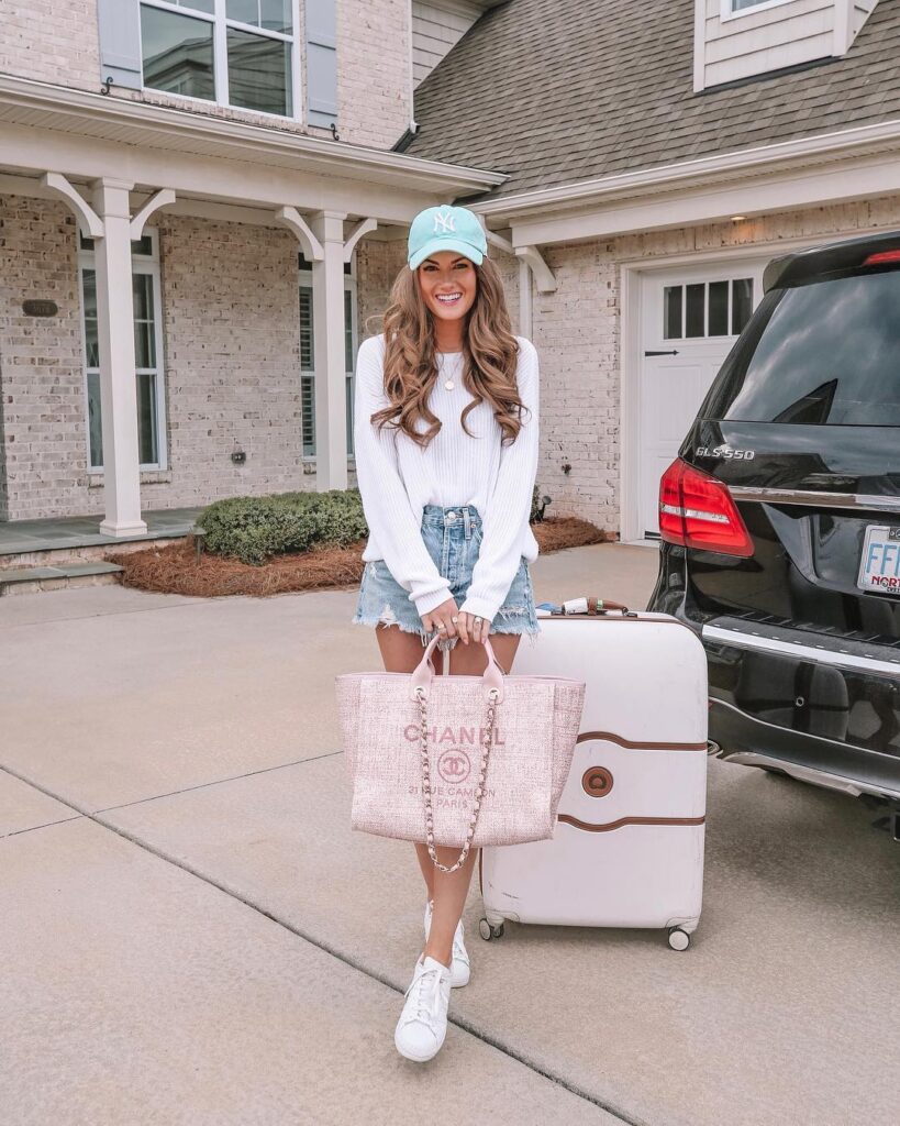 Woman wearing a blue cap and white sweater, smiling outside a house with a Chanel tote and suitcase ready for travel.