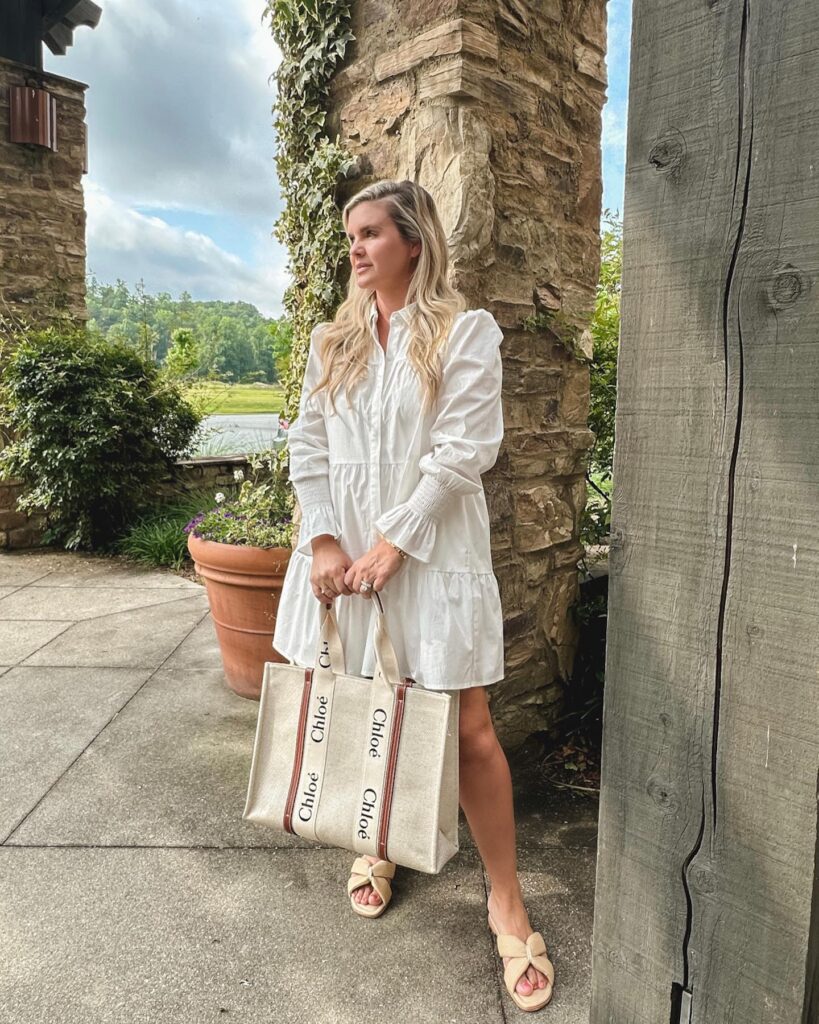 Woman in a stylish white dress poses with a Chloé tote bag beside a stone wall and lush greenery outdoors.
