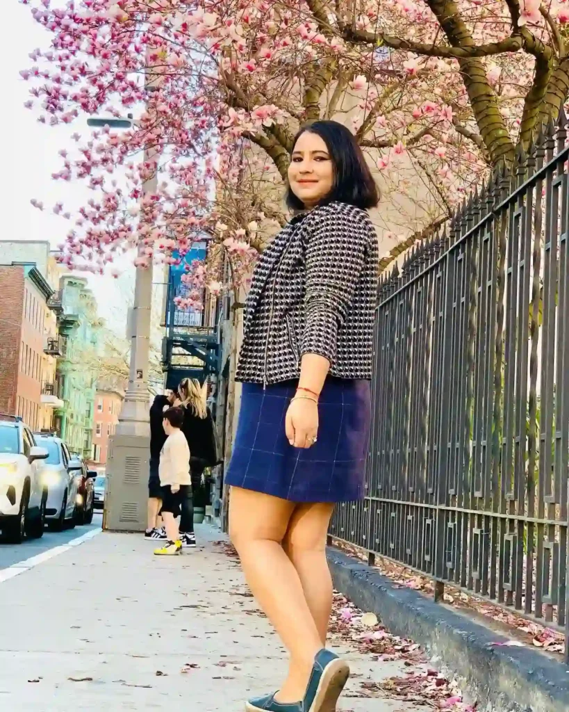Young woman in stylish outfit poses near blooming cherry blossom trees on a city street, capturing spring's beauty.