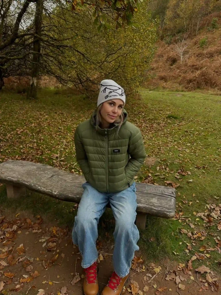 Woman in a green puffer jacket and beanie sitting on a wooden bench in a scenic autumn landscape.