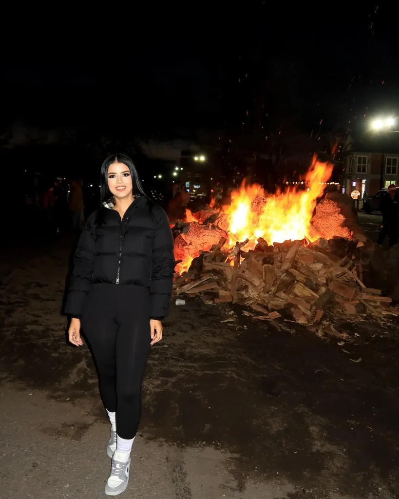 Woman in a black puffer jacket poses near a large bonfire at night, surrounded by a festive atmosphere.